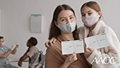A masked, female patient puts her arm around another masked, female patient and they both hold up vaccination cards