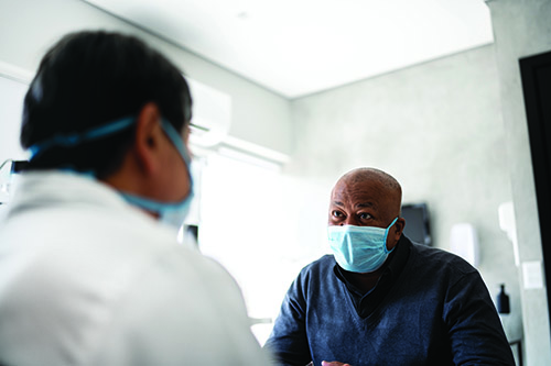 A  male patient in a mask speaking to a health professional who is also wearing a mask