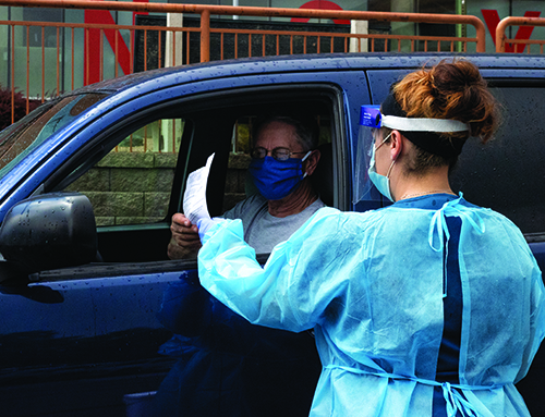 A woman in a gown and mask handing a paper to a man in a car.