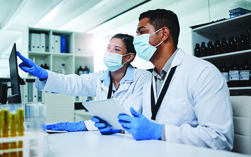 A woman laboratorian and male laboratorian in lab coats, masks, and gloves, sitting at a table in a lab and looking at a screen.