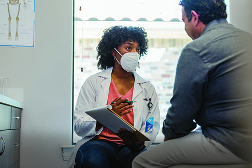 A woman doctor wearing a mask while sitting down and speaking to a male patient