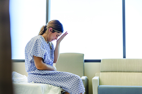 A woman patient in a hospital gown sitting on the edge of the bed putting her hand to her head