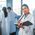 A female lab professional looking at the camera holds a notebook in her arm. In the background other lab professionals with their backs to the camera confer.