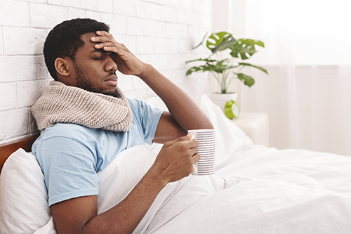 An African American male lying in bed while holding a mug in one hand and putting his other hand over his face.
