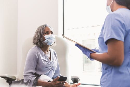 A woman wearing a mask sits and looks up at a nurse.